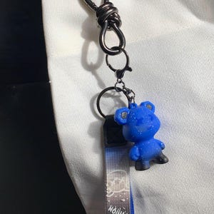 May include: A blue bear keychain with a black and white strap that says "Fashion". The keychain is attached to a silver clasp. The background is a white tote bag.