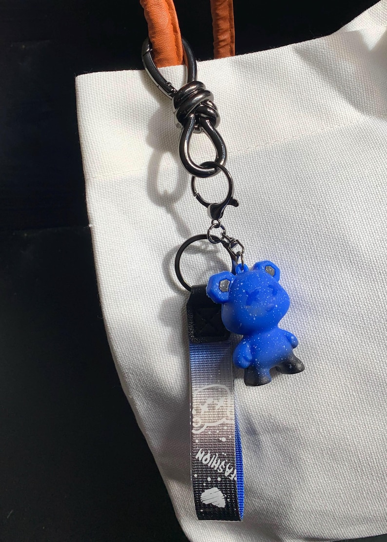 May include: A blue bear keychain with a black and white strap that says "Fashion". The keychain is attached to a silver clasp. The background is a white tote bag.