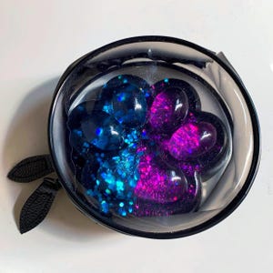 May include: A round, clear zippered pouch containing a resin paw print ornament. The paw print features a split color design with blue glitter on one side and pink glitter on the other, set against a dark base with a glossy, smooth finish.