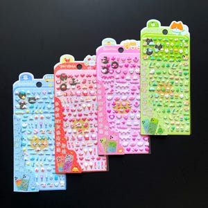 May include: Four vertical sheets of small, puffy stickers in blue, red, pink, and green. Each sheet features various miniature icons like food, animals, and shapes, with Chinese text and illustrations of decorated mobile phones at the bottom.