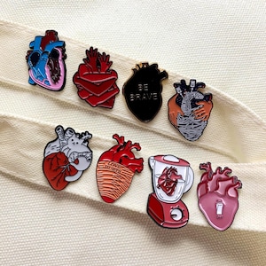 May include: A collection of enamel pins with heart designs. The pins feature anatomical hearts in various colors, including blue, red, and black. One pin has the text "BE BRAVE", and another says "LOVE HURTS". A blender pin and a pin with an on/off switch are also included.