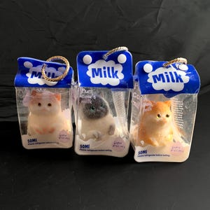 May include: Three clear plastic containers shaped like milk cartons, each containing a small cat figurine. The cartons feature blue tops with the text Milk, 50ml, and Super delicious. The cats are white, grey, and orange, each with a gold hanging loop.
