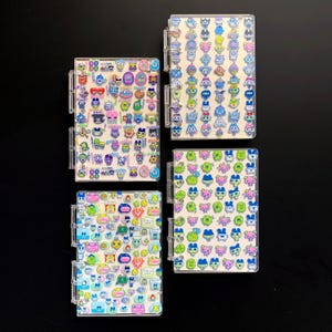 May include: Four clear, plastic-covered notebooks, each filled with colorful cartoon character stickers. The stickers feature various shapes and colors, including blue, green, pink, and yellow. The notebooks have a binder-style spine.
