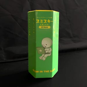 May include: A hexagonal green cardboard box featuring a cartoon character sitting and using a laptop. The text reads Smiski Work and Glow in the Dark in yellow lettering. The box has Japanese characters printed vertically along the side panels.