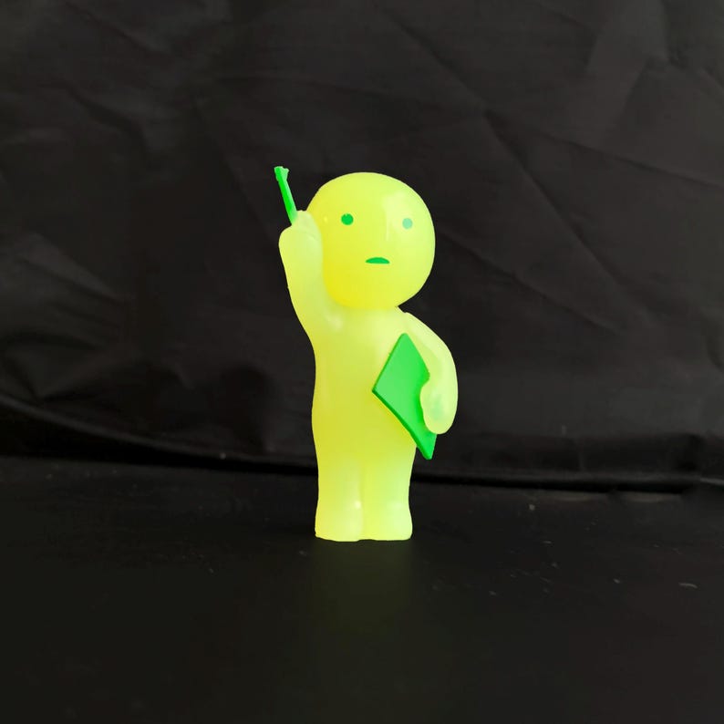 May include: A small, translucent neon yellow figurine shaped like a stylised person. The figure has a round head with two green dots for eyes and a green line for a mouth. It holds a green rectangular object in one hand and raises the other arm.