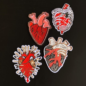 May include: Four embroidered patches featuring anatomical heart designs. One is a realistic red heart, one shows a heart within a ribcage, one is a red heart pierced by multiple needles, and one is a dark heart with prominent white vessels.