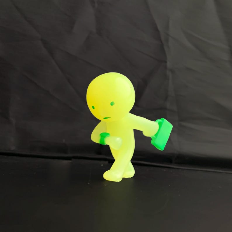May include: A small, translucent neon yellow figurine of a person walking. The figure has a round head with two green dots for eyes and a green line for a mouth. It wears a green watch on its wrist and carries a green rectangular briefcase.
