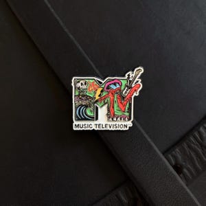 May include: A metal enamel pin featuring the MTV logo with a colorful graphic design. The logo includes a skull, a blue wave, a lightning bolt, and a guitar. The text MUSIC TELEVISION is printed in black capital letters at the bottom of the pin.