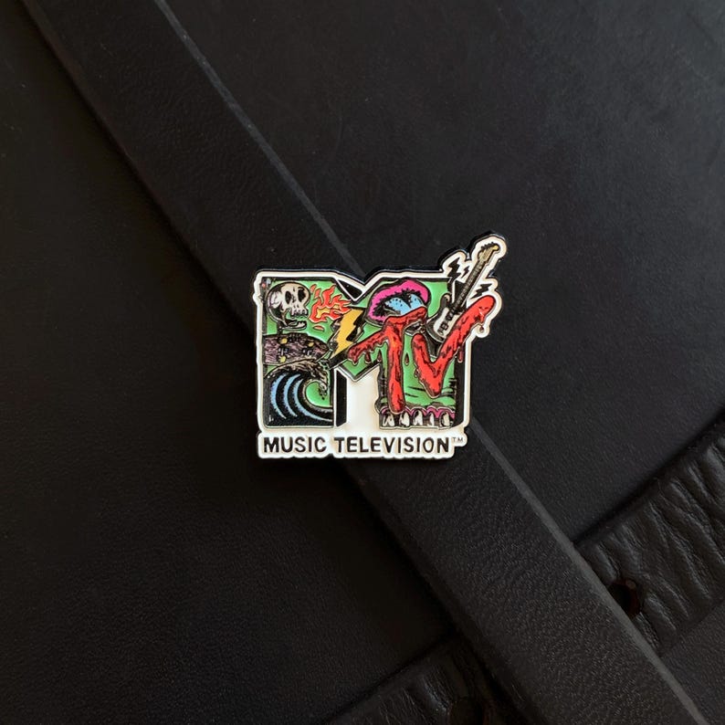 May include: A metal enamel pin featuring the MTV logo with a colorful graphic design. The logo includes a skull, a blue wave, a lightning bolt, and a guitar. The text MUSIC TELEVISION is printed in black capital letters at the bottom of the pin.