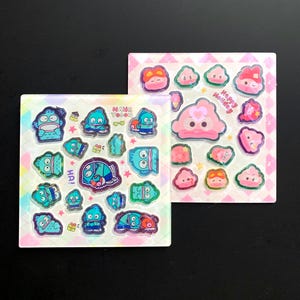 May include: Two square sticker sheets featuring cartoon characters. The left sheet has blue fish-like creatures with the text HANG YODON. The right sheet has pink blob-like characters with the text Happy Holiday. Both sheets include small stars.