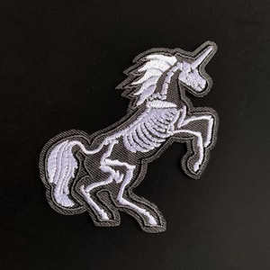 May include: An embroidered patch featuring a white skeleton of a unicorn on a black background. The design shows the unicorn rearing up with its mane, tail, and horn clearly outlined in white thread against the dark fabric of the patch.