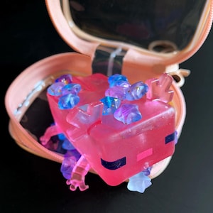 May include: A translucent pink axolotl figurine with dark blue eyes and a pink mouth, adorned with small blue and purple star-shaped beads on its head and body, resting inside a small, open, peach-colored zippered carrying case.