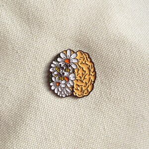 May include: A gold-colored brain-shaped enamel pin with a floral design. The left side of the pin features white daisies with orange centers, while the right side depicts the brain's structure. The pin is set against a textured, off-white background.
