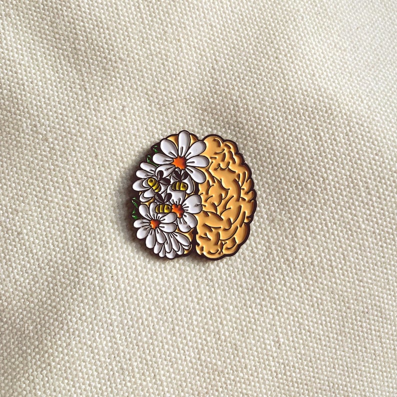 May include: A gold-colored brain-shaped enamel pin with a floral design. The left side of the pin features white daisies with orange centers, while the right side depicts the brain's structure. The pin is set against a textured, off-white background.