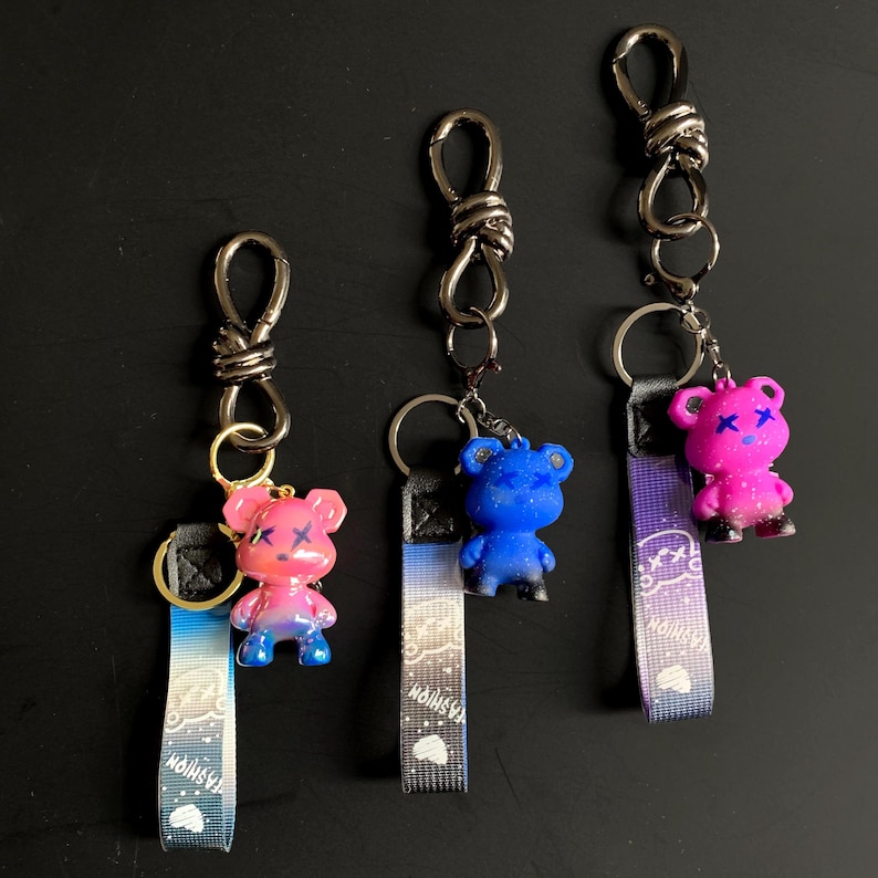 May include: Three keychains with bear-shaped charms. The charms are pink, blue, and pink with black accents. Each keychain has a metal clasp and a fabric strap with the word "FASHION" printed on it.