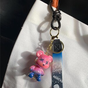 May include: A pink and blue bear-shaped keychain with black cross-shaped markings on its face. The keychain has a black and gold clasp, a blue and white patterned strap with the word "FASHION", and a black and orange handle.