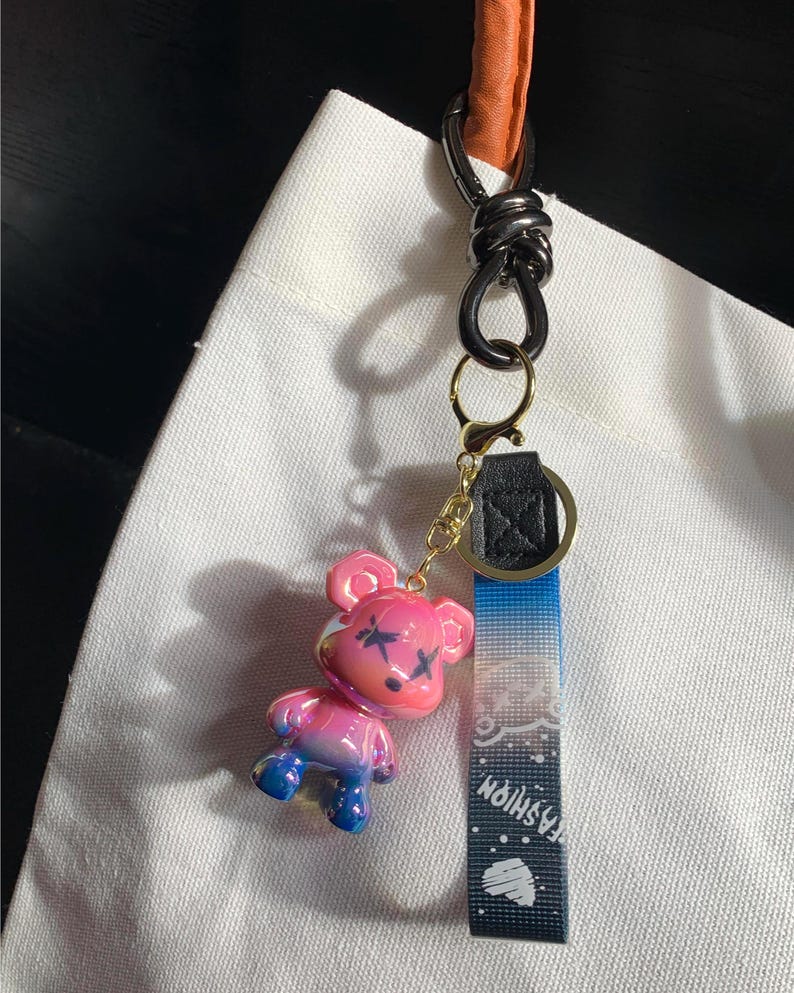 May include: A pink and blue bear-shaped keychain with black cross-shaped markings on its face. The keychain has a black and gold clasp, a blue and white patterned strap with the word "FASHION", and a black and orange handle.