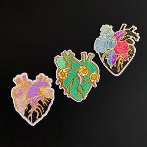 May include: Three embroidered patches shaped like anatomical hearts. The left patch is purple and dark blue with gold stitching. The center patch is green with yellow flowers. The right patch features a black heart with blue and pink floral accents.