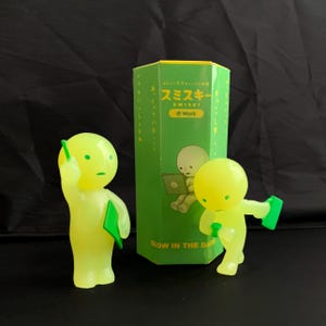 May include: Two glow-in-the-dark Smiski figures in lime green, one standing with a green folder and one leaning forward with a green folder, positioned next to a hexagonal green box featuring the text Smiski Work and a graphic of a figure.