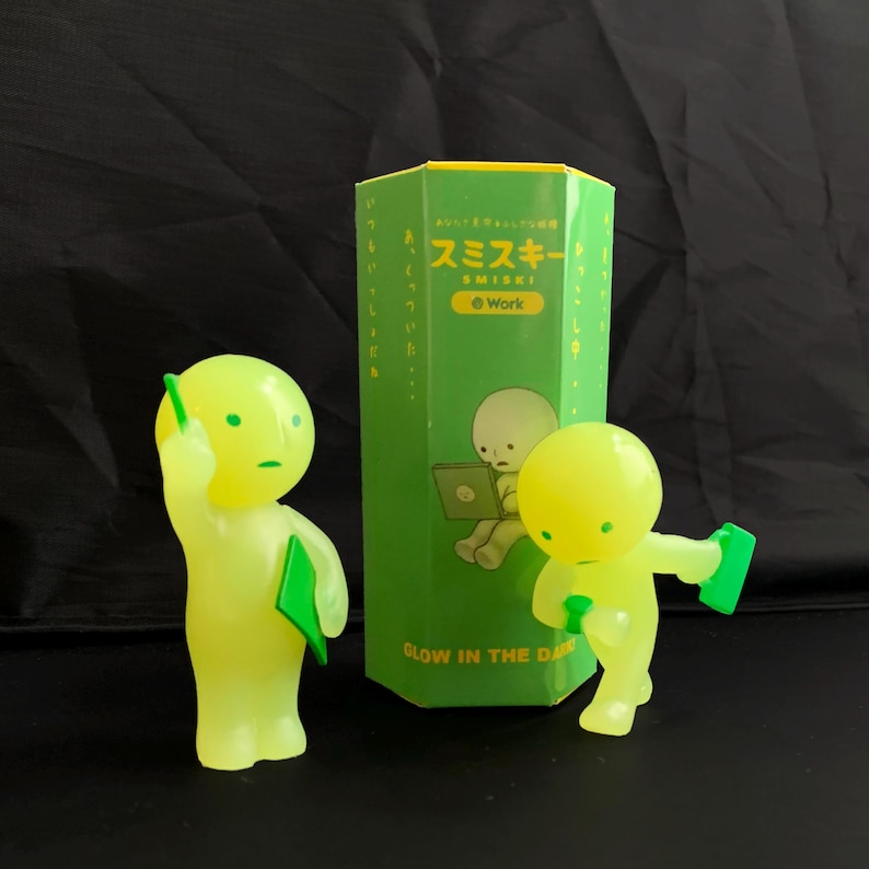 May include: Two glow-in-the-dark Smiski figures in lime green, one standing with a green folder and one leaning forward with a green folder, positioned next to a hexagonal green box featuring the text Smiski Work and a graphic of a figure.