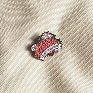 May include: A pink brain-shaped enamel pin with a white banner that reads "BE KIND TO YOUR MIND." The pin is surrounded by flowers and leaves. The pin is on a cream-colored fabric.