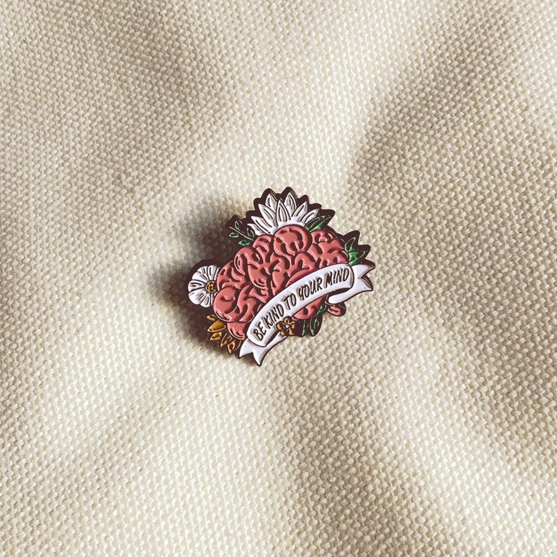May include: A pink brain-shaped enamel pin with a white banner that reads "BE KIND TO YOUR MIND." The pin is surrounded by flowers and leaves. The pin is on a cream-colored fabric.