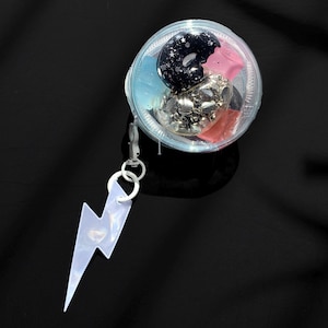 May include: A clear, round container with a zipper, holding a black donut-shaped charm with white stars, a pink star, and silver charms. A white lightning bolt charm hangs from the zipper.