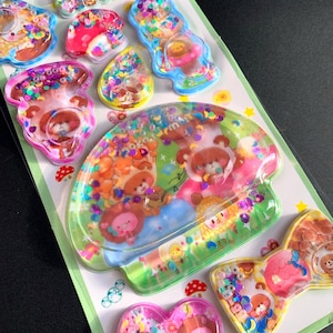 May include: A sheet of colorful resin stickers with glitter and cartoon bear designs. The stickers are various shapes, including a heart, a bow, and a mushroom. The stickers are filled with colorful confetti and glitter. The words "glitter magic" are visible in the top left corner.