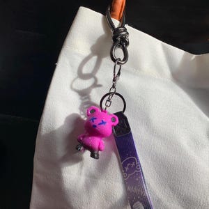 May include: A pink bear keychain with blue accents and a purple strap. The bear has a cross design on its face. The strap has the word "Fashion" and a graphic design. The keychain is attached to a silver metal clasp.