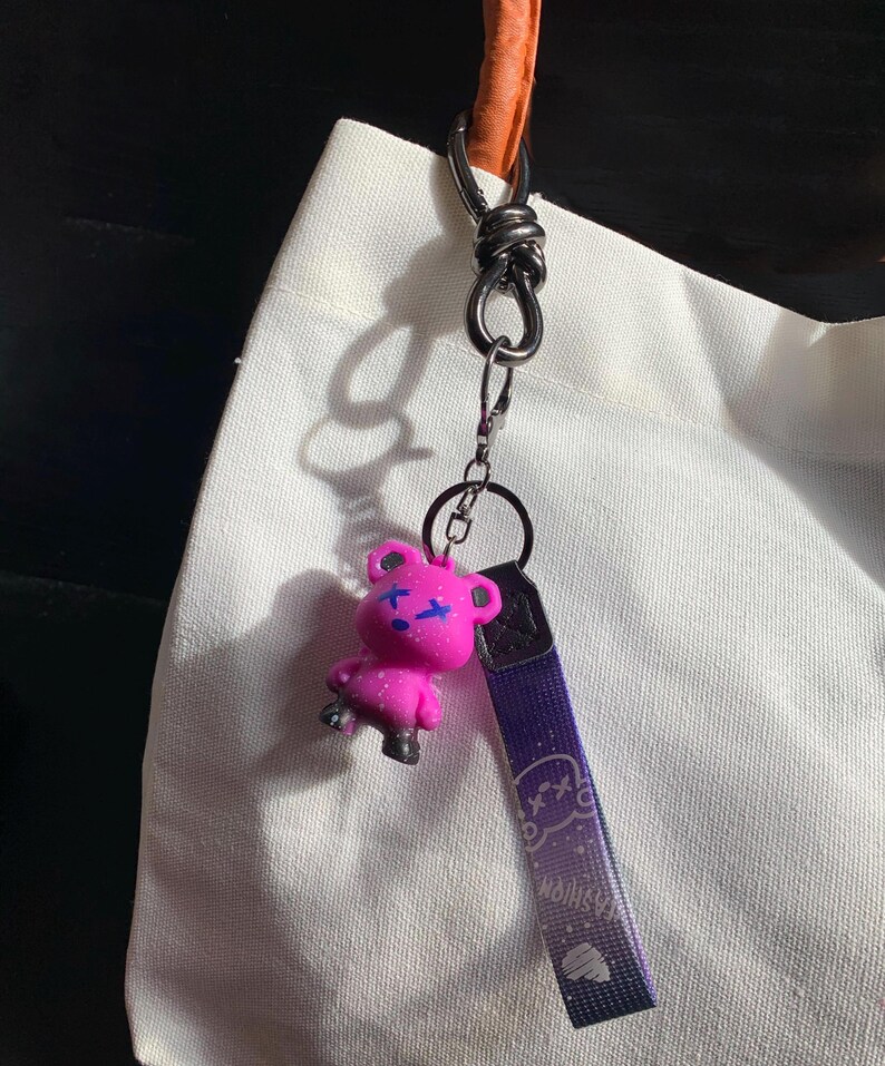 May include: A pink bear keychain with blue accents and a purple strap. The bear has a cross design on its face. The strap has the word "Fashion" and a graphic design. The keychain is attached to a silver metal clasp.