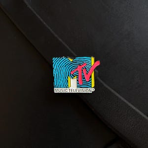 May include: A rectangular enamel pin featuring the MTV logo with a bright blue fingerprint pattern on the letter M, a bold pink TV lettering overlay, and the words MUSIC TELEVISION printed in black on a white rectangular base at the bottom.