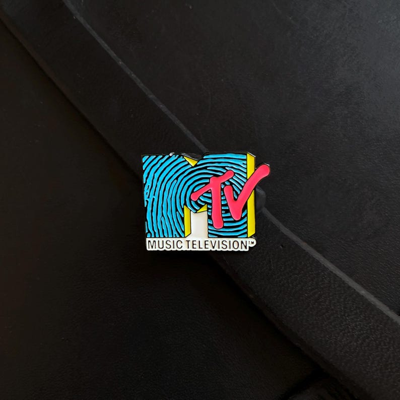 May include: A rectangular enamel pin featuring the MTV logo with a bright blue fingerprint pattern on the letter M, a bold pink TV lettering overlay, and the words MUSIC TELEVISION printed in black on a white rectangular base at the bottom.