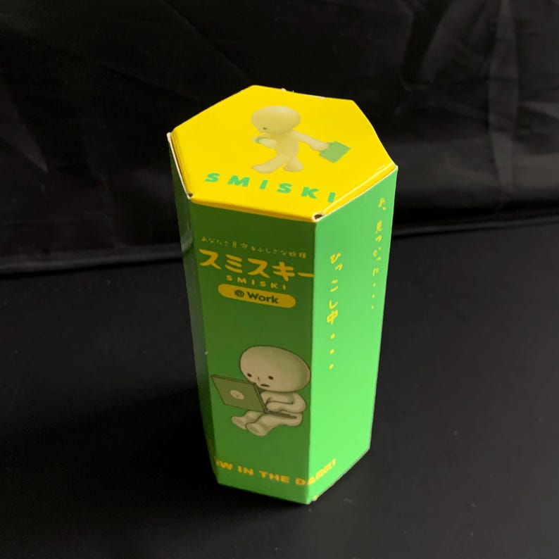 May include: A hexagonal cardboard box in bright green and yellow featuring the Smiski Work character. The top shows a yellow Smiski figure with a briefcase. The side displays a Smiski figure using a laptop with the text Smiski Work and Glow in the Dark.