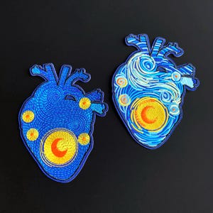May include: Two embroidered patches shaped like anatomical hearts. The patches feature a blue background with swirling patterns, yellow circular sun or moon motifs, and orange crescent details, all rendered in dense, colorful thread work.