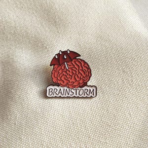 May include: A red brain-shaped enamel pin with a red umbrella and the word "BRAINSTORM" in white lettering. The pin is set against a neutral, textured background.