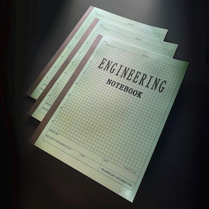 May include: Three engineering notebooks with light green graph paper and brown spines are stacked on a dark surface. The cover of the top notebook reads "ENGINEERING NOTEBOOK" in black text. The notebooks have a grid pattern and areas for titles and signatures.