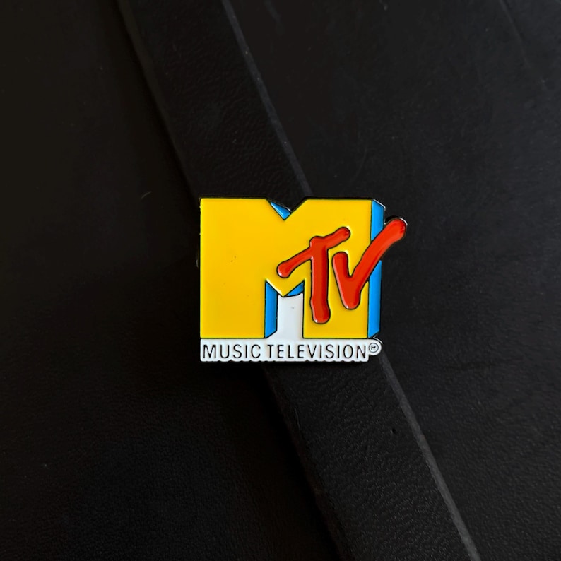 May include: A metal enamel pin featuring the MTV logo with a large yellow letter M, a red TV script overlay, and the text MUSIC TELEVISION in white capital letters at the bottom. The design includes blue accents on the side of the letter M.