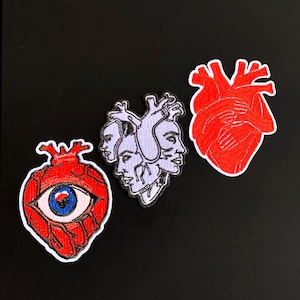 May include: Three embroidered patches featuring anatomical designs. One patch is a red heart with a large blue eye in the center. The middle patch is a light purple cluster of human faces. The third patch is a red heart with hands wrapped around it.