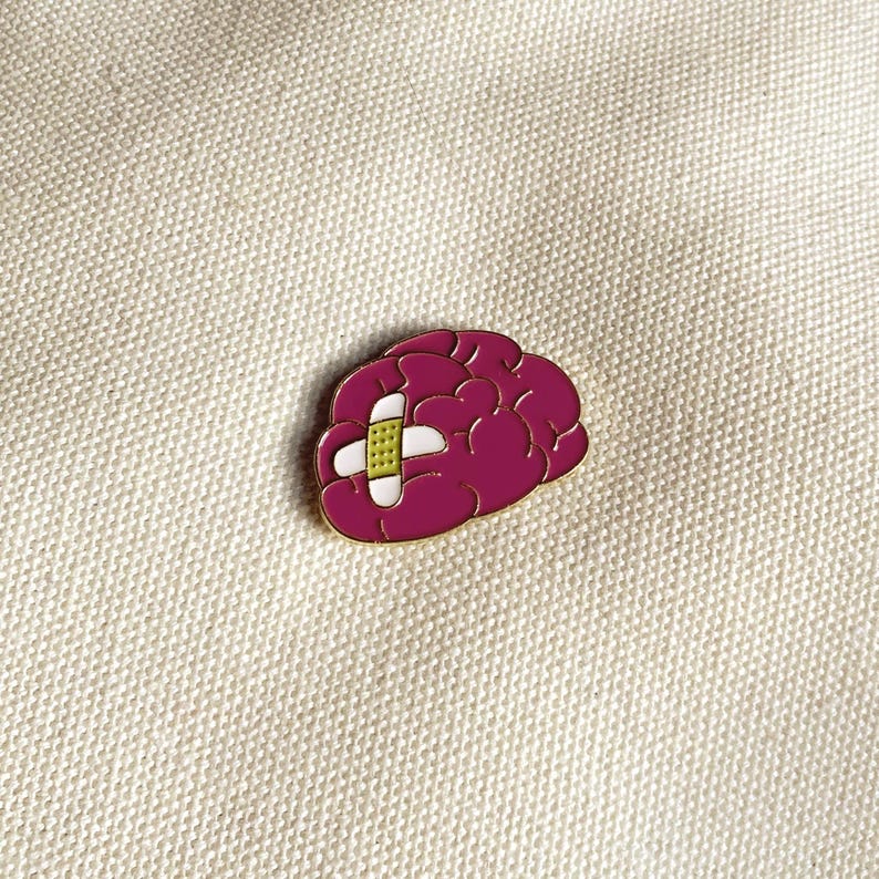 May include: A magenta-colored brain-shaped enamel pin with a white and yellow bandage across the center. The pin has a gold-colored outline and is set against a cream-colored fabric background.