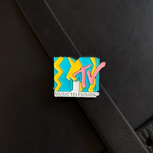 May include: A rectangular enamel pin featuring the MTV logo with a bright blue and yellow zigzag pattern. The letters TV are in pink, and the text MUSIC TELEVISION is printed in white on a black background at the bottom of the pin design.
