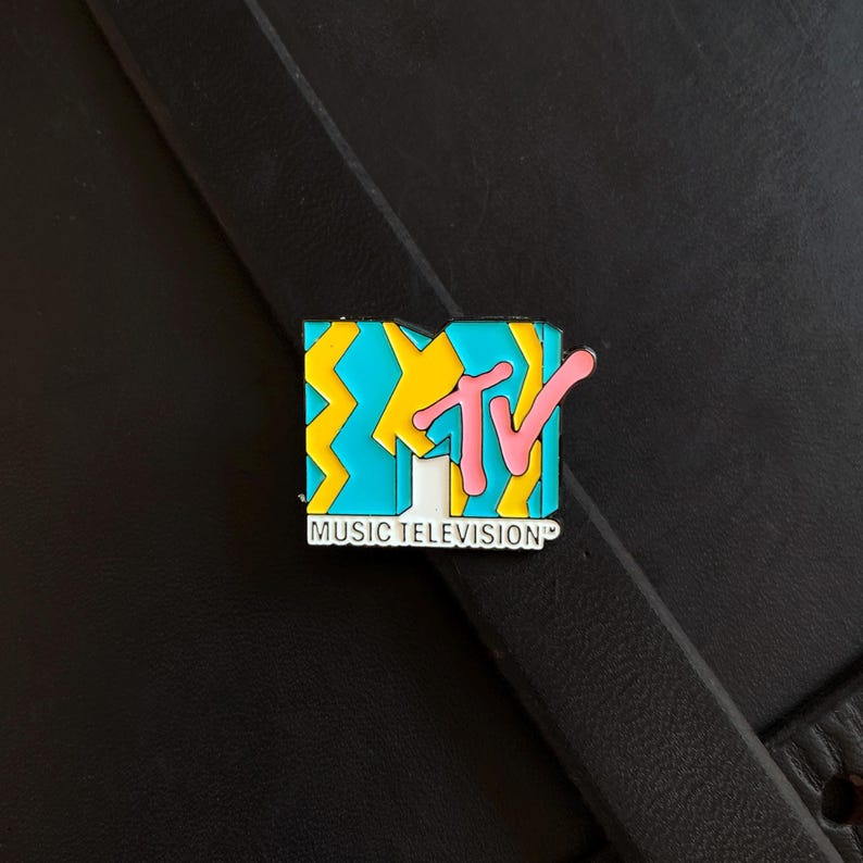 May include: A rectangular enamel pin featuring the MTV logo with a bright blue and yellow zigzag pattern. The letters TV are in pink, and the text MUSIC TELEVISION is printed in white on a black background at the bottom of the pin design.