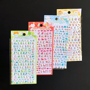 May include: Four vertical sheets of small, puffy stickers featuring various colorful icons. The sheets include themes of green nature elements, pink fruit and hearts, blue travel motifs, and yellow party items, each with Japanese text at the top.