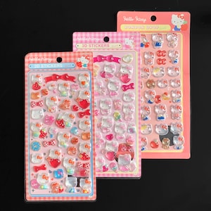 May include: Three packs of 3D stickers featuring Hello Kitty characters in various poses and designs. The stickers are in clear, pink, and red colors, with bows, hearts, and other decorative elements. The packaging includes the text "Hello Kitty" and "3D Stickers".