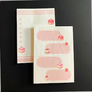 May include: Two pink and white notepads with a dessert theme. One notepad has a list of pink hearts and cake illustrations. The other notepad has pink ovals for writing and cake and strawberry illustrations. The word "plan" is at the top of one notepad.