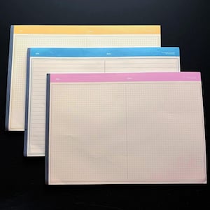 May include: Three notepads with grid paper and colored borders in yellow, blue, and pink. Each notepad has a different color border and a lined section for the date. The notepads are stacked on top of each other.