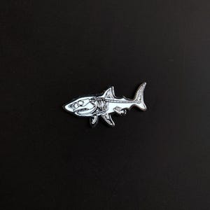 May include: A silver-toned metal enamel pin shaped like a shark. The design features a detailed black and white skeletal anatomy of the shark, showcasing the skull, rib cage, and spine structure within the silhouette of the fish body.