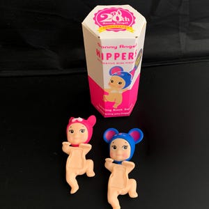 May include: Two Sonny Angel Hippers decorative mini figures, one in a pink mouse hat and one in a blue mouse hat, positioned in front of a hexagonal white and pink box labelled 20th Anniversary Sonny Angel HIPPERS Looking Back Series.
