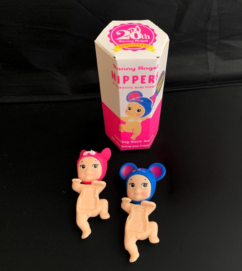 May include: Two Sonny Angel Hippers decorative mini figures, one in a pink mouse hat and one in a blue mouse hat, positioned in front of a hexagonal white and pink box labelled 20th Anniversary Sonny Angel HIPPERS Looking Back Series.