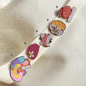 May include: A collection of five brain-themed enamel pins. The pins feature various designs, including a brain with a bandage, a brain with flowers, and a brain with the word "Brainstorm". The pins are colorful and detailed, with a variety of shapes and sizes.