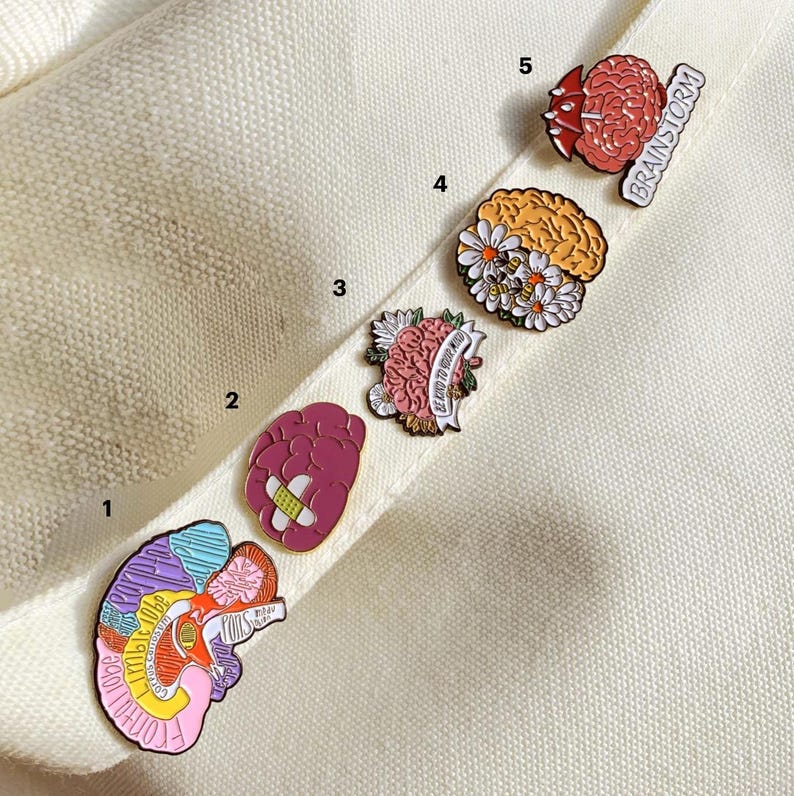 May include: A collection of five brain-themed enamel pins. The pins feature various designs, including a brain with a bandage, a brain with flowers, and a brain with the word "Brainstorm". The pins are colorful and detailed, with a variety of shapes and sizes.